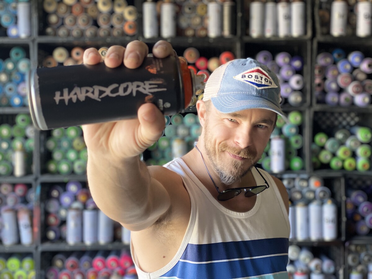 Man in a Boston cap holding a black spray paint can labeled Hardcore in front of a wall stocked with spray paint cans